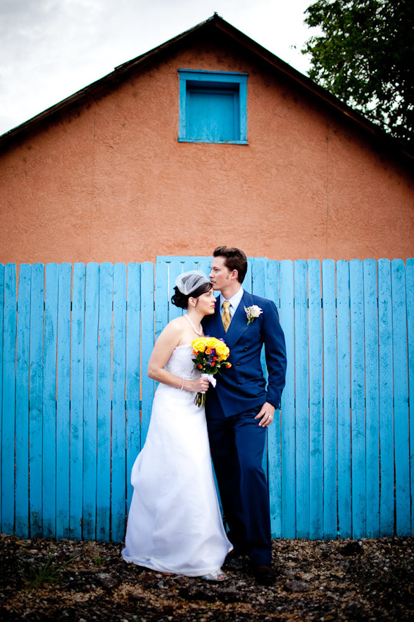 photo of the bride and groom standing in front of a bright aqua colored fence and orange building - bride is wearing a white a-line style dress and birdcage veil while holding a yellow, red, and green bouquet and groom is wearing blue suit - photo by New Mexico based wedding photographers Twin Lens